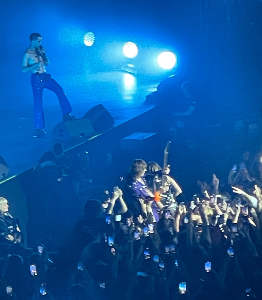 A picture of band members from Maneskin, the singer is on the state in blue light, the guitarist and bassist are playing in the sea of people below