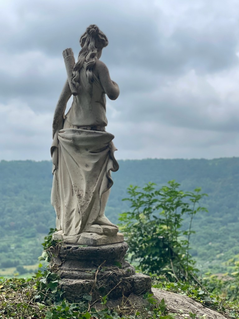 A stone statue of Diana from behind. You see her chiton draped and a quiver for arrows on her back. She seems to be looking down the slope of the hill towards the lake.