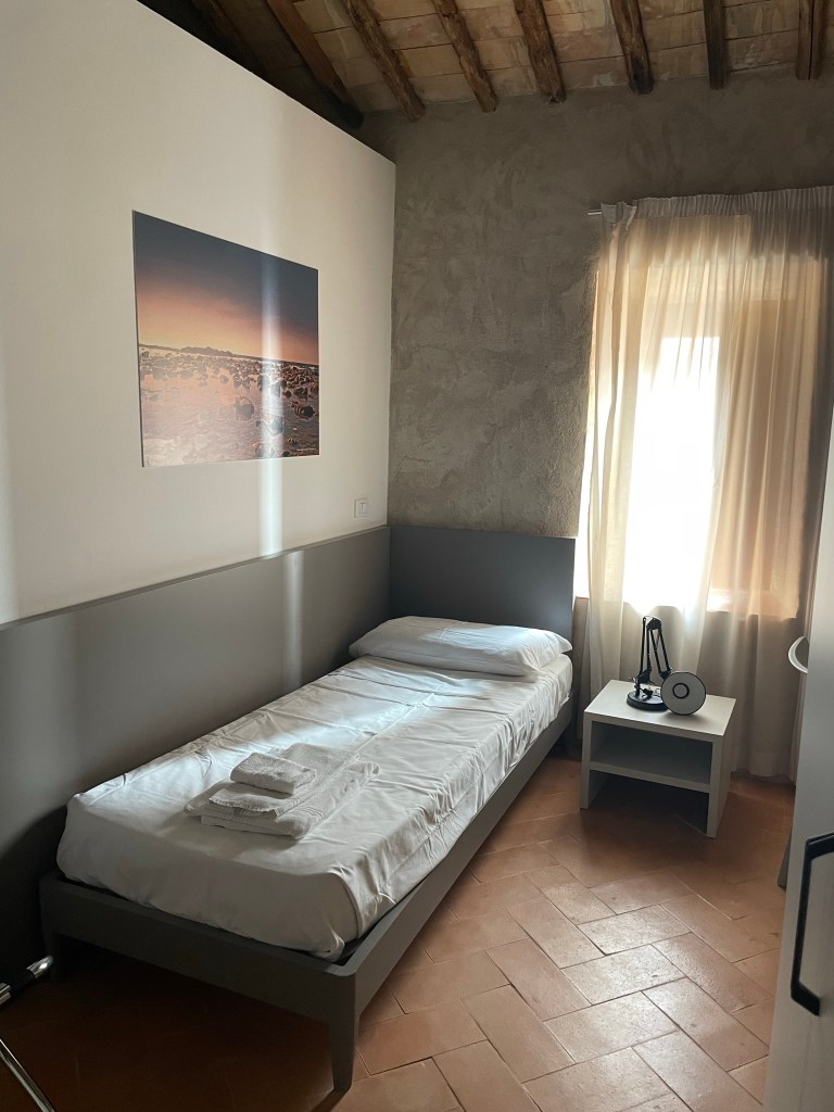 A single bed with white sheets in a small hotel room with terracotta tile floor.