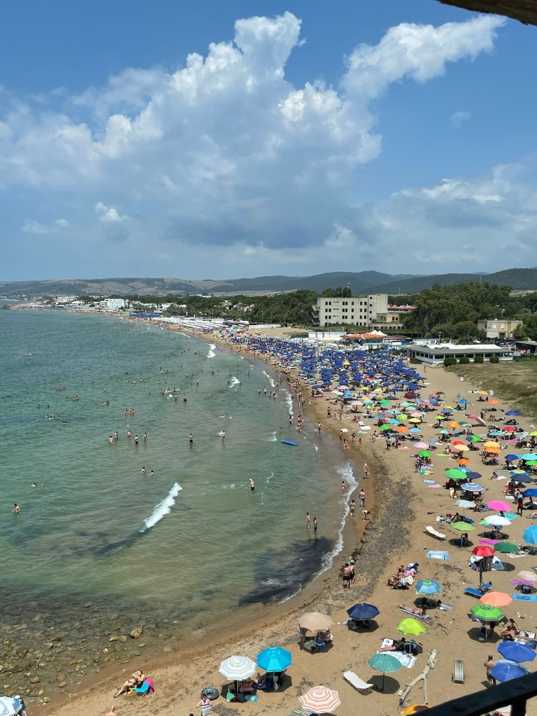 View of the beach from inside the castle, there's lots of umbrellas on the sand next to azure water and a blue sky.
