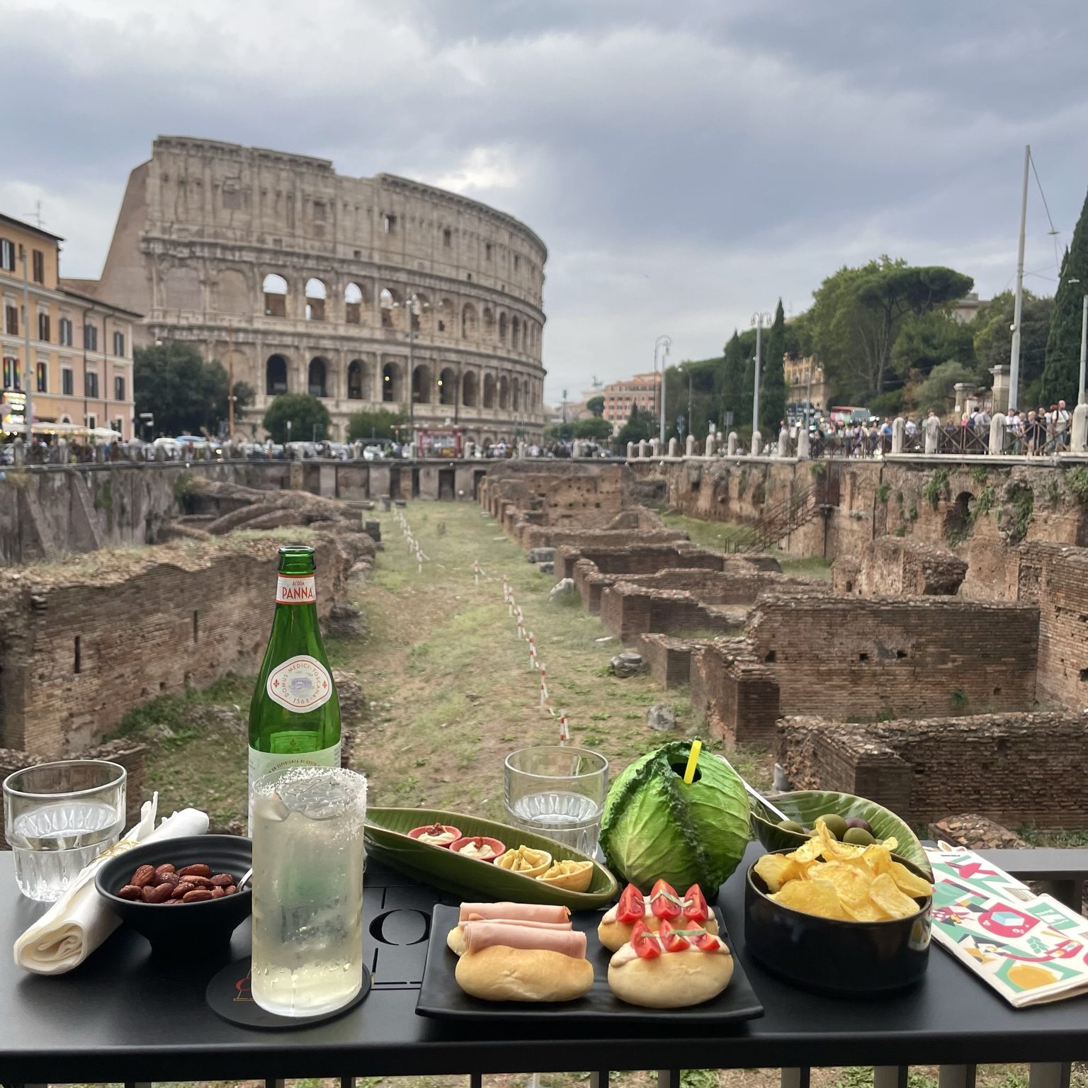 A view of the Ludus Magnus from The Court, a fantastic bar with creative drinks and great appetizers. The Colosseum is in the background, the ruins of the Ludus Magnus in the middle, and in the foreground is an assortment of drinks and snacks. 