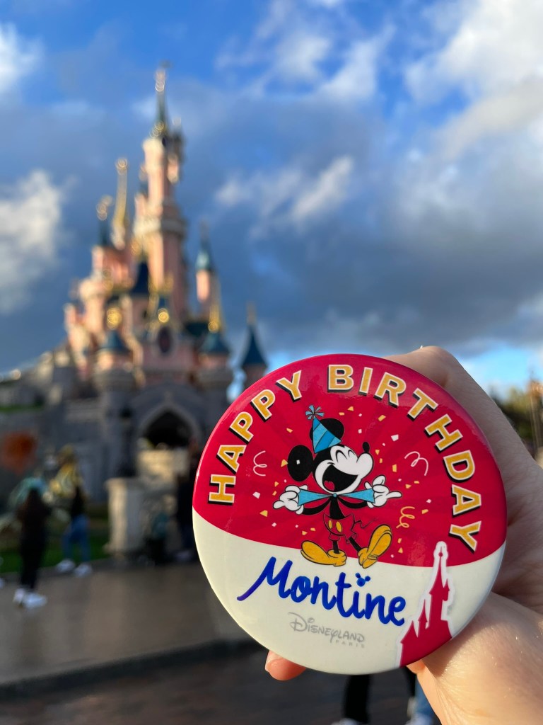 My hand holding up my birthday pin, with Disneyland Paris’ Sleeping Beauty Castle in the background. 